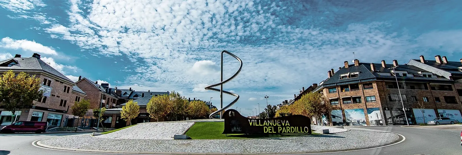 Glorieta de Villanueva del Pardillo con la escultura Rastro Orbital y el rótulo del pueblo, rodeada de bloques de ladrillo